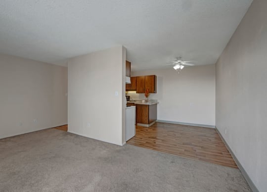 Spacious, empty living room here at Cedar Gardens Apartment Homes with beige walls, carpeted floor, and a separate area with wood flooring. A small kitchen with wooden cabinets and ceiling fan.