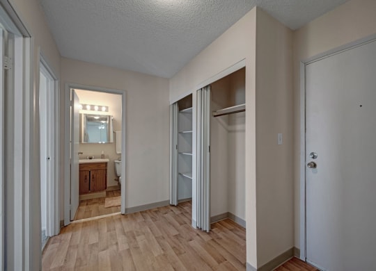 Minimalist hallway here at Cedar Gardens Apartment Homes with light wood flooring. Open closet with sliding doors on the right. Bathroom with mirror and lights visible in the background.