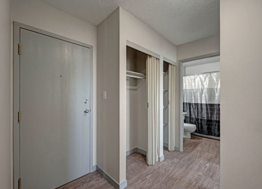Compact hallway here at Cedar Gardens Apartment Homes with a light gray entry door on the left, adjacent closet with sliding doors, and a glimpse into a bathroom with a white and black shower curtain.