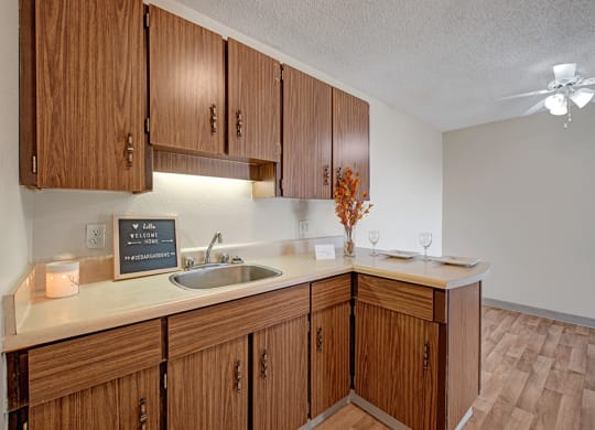 Cozy kitchen here at Cedar Gardens Apartment Homes with wooden cabinets, a small sink, and a lighted backsplash. A welcoming sign, candle, and two place settings add warmth.