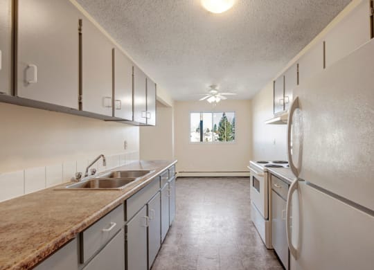 Narrow kitchen here at Cedar Gardens Apartment Homes with light gray cabinets, white appliances, and granite countertops. A window at the end overlooks trees, creating an airy atmosphere.
