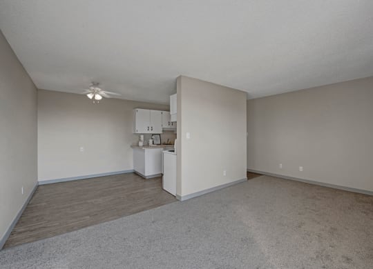 Spacious, empty apartment here at Cedar Gardens Apartment Homes with beige walls and carpeted floors. A small kitchen area with white cabinets and a ceiling fan is visible, creating a neutral tone.