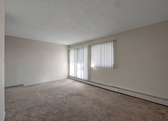 Empty room here at Cedar Gardens Apartment Homes with beige carpet and white walls, featuring two vertical blind-covered windows letting in soft light, creating a calm, minimalist atmosphere.
