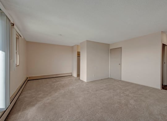 An empty room here at Cedar Gardens Apartment Homes with beige walls and carpet, featuring a closed door, small window on the left, and open closet. Soft lighting creates a calm atmosphere.