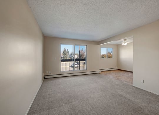 Spacious, empty living room here at Cedar Gardens Apartment Homes with beige walls and carpet, large window showing trees outside. Adjacent dining area with a ceiling fan and tiled floor.
