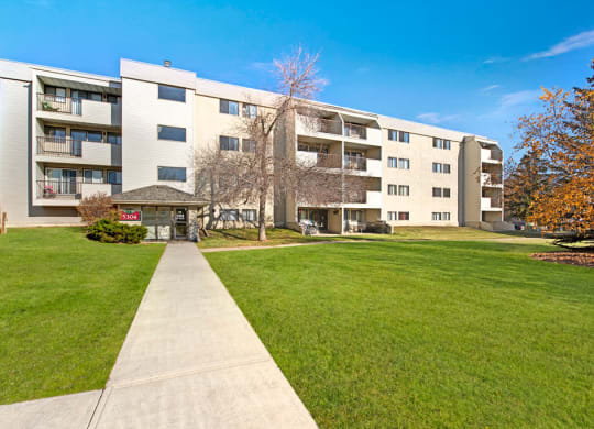 Four-story Cedar Gardens Apartment Homes building with balconies and a lawn, under a clear blue sky. A pathway leads to the entrance, creating a welcoming atmosphere.