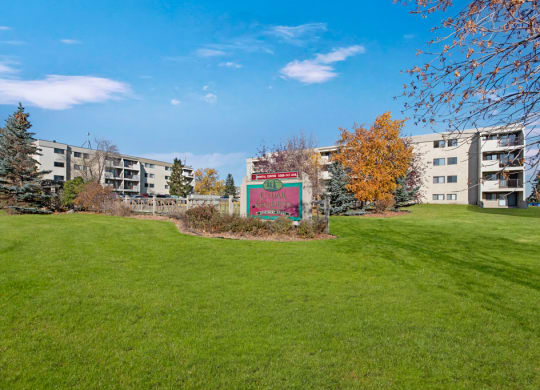 Expansive grassy lawn with a sign for “Cedar Gardens” centered between two beige apartment buildings. Bright autumn trees under a clear blue sky.