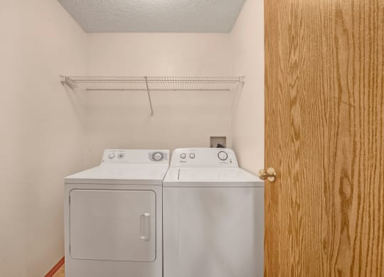 Laundry room here at Edgewood Estates Apartment Homes with a white washer and dryer side by side under a wire shelf on a beige wall. A wooden door is partially open on the right.