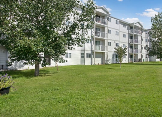 Four-story white Edgewood Estates Apartment Homes building with balconies, surrounded by lush green grass and trees, under a clear blue sky. Calm and inviting atmosphere.