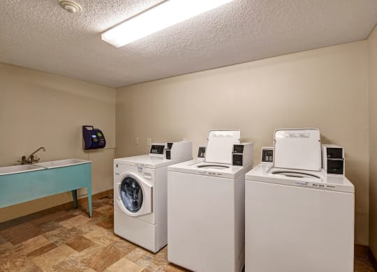 Laundry room here at Lakewood Heights Apartment Homes with two white top-load washing machines and a front-load dryer. A utility sink with a faucet is on the left. Beige walls and tiled floor.