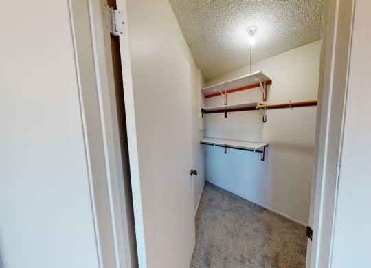 A small walk-in closet here at Lakewood Heights Apartment Homes with beige carpet, white walls, and a single ceiling light. Two wooden rods and white shelves for storage are visible.