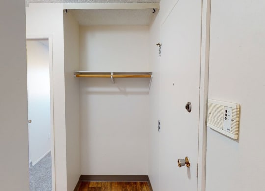 Empty white closet here at Lakewood Heights Apartment Homes with a wooden rod and shelf, adjacent to a door with a round knob. A light switch and intercom are mounted on the wall.