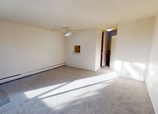 Empty bedroom here at Lakewood Heights Apartment Homes with beige walls and gray carpet, illuminated by sunlight. Features include a ceiling fan, small wall cutout, and an open door. Minimalist and bright atmosphere.
