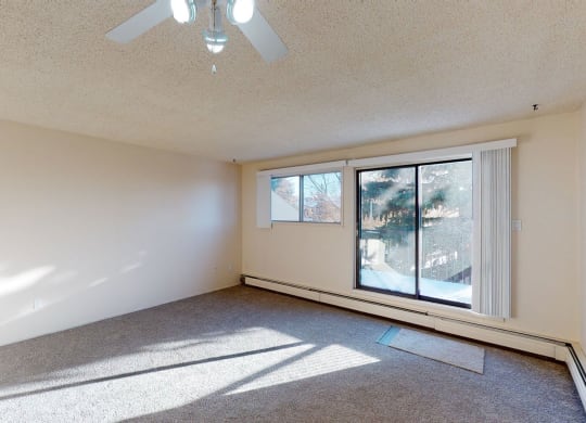 Empty room here at Lakewood Heights Apartment Homes with beige walls, carpeted floor, and ceiling fan. Large sliding glass doors and a window let in sunlight, casting shadows across the room.