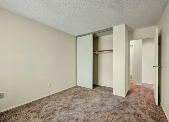 Empty bedroom here at Lakewood Heights Apartment Homes with beige walls and carpet, featuring an open closet with sliding doors. A partially open door reveals a hallway, creating a calm, neutral tone.