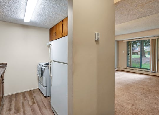Compact kitchen here at Lakewood Heights Apartment Homes with wood cabinets and a white stove, adjacent to a bright living room with beige carpet and large window showing a grassy view. Cozy atmosphere.