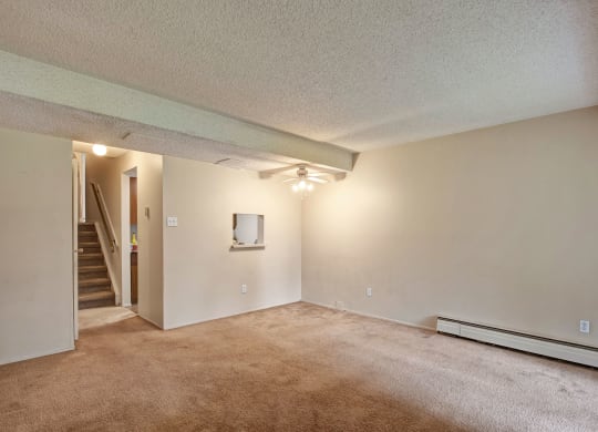 Empty living room here at Lakewood Heights Apartment Homes with beige walls and carpet, ceiling fan, and a small window opening to a kitchen area. Stairs are visible in the background.