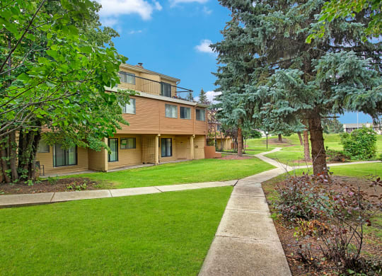 Spacious green backyard here at Lakewood Heights Apartment Homes with a beige two-story house, surrounded by lush trees and manicured lawns. A winding concrete path leads through the serene landscape.