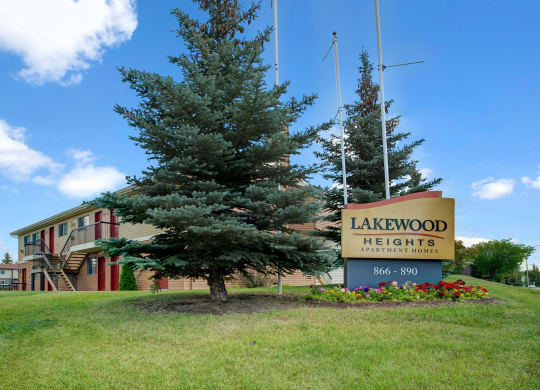 Sign for Lakewood Heights Apartment Homes set on a manicured lawn with colorful flowers and tall evergreen trees. A clear, sunny sky above.