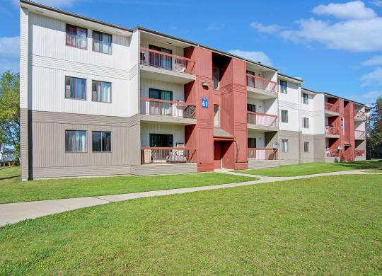 Three-story Madison Park Apartment Homes building featuring red and beige panels, balconies, and a green lawn. A clear blue sky suggests a sunny day.
