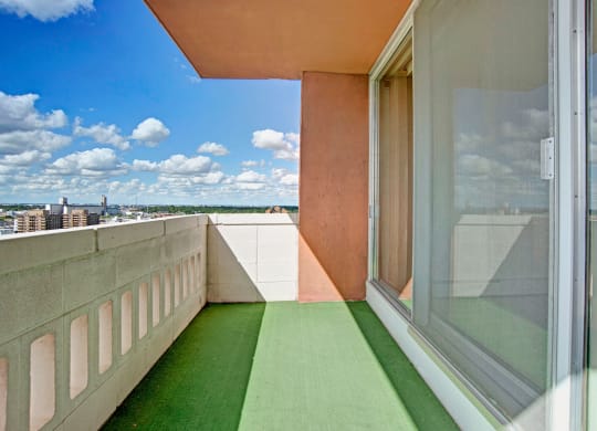 Sunny balcony here at Marquis Towers Apartment Homes with green artificial grass flooring and white railing, offering a panoramic city view under a blue sky with fluffy clouds.