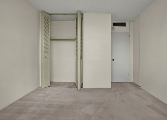 Empty bedroom here at Marquis Towers Apartment Homes with beige walls and carpet. Open closet with light green louvered doors on the left. Closed white door with vent above on the right. Minimalist, calm atmosphere.