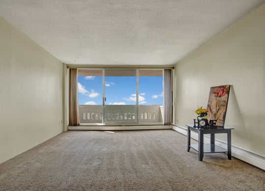 Empty room here at Marquis Towers Apartment Homes with beige carpet and walls, featuring a large glass door opening to a balcony. A small table with a floral picture and "Home" decor sits to the right.