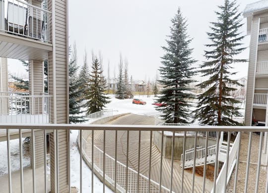 View from a balcony here at The Pointe at Applewood overlooking snow-covered ground, flanked by tall, snow-dusted evergreens. A red car is parked below, near beige apartment buildings. Winter ambiance.