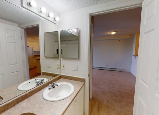 Bathroom here at The Pointe at Applewood with beige countertop, sink, and mirror. Bright lights above. Open door reveals a kitchen with white cabinets and an adjacent room. Cozy and inviting.
