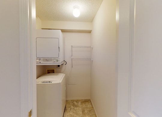 Compact laundry room here at The Pointe at Applewood with a stackable washer and dryer, beige walls, and a bright overhead light. Two wire shelves are mounted on the right wall.