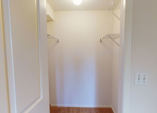 Empty closet here at The Pointe at Applewood with beige walls and a beige carpeted floor, illuminated by a ceiling light. Metal racks are mounted on the left and right sides.