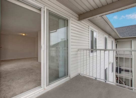 View of a small balcony here at The Pointe at Applewood with white railing attached to a white building, leading to an empty room with beige carpet and a ceiling light.