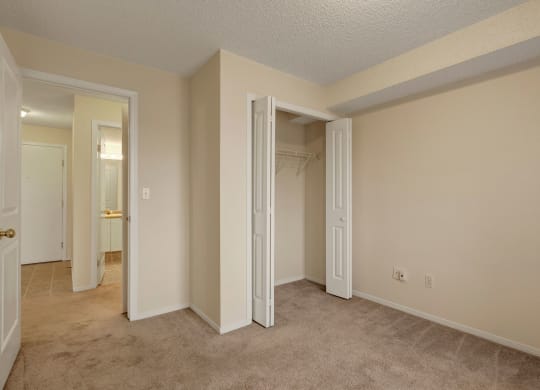 Empty beige room here at The Pointe at Applewood with a carpeted floor, open closet with white folding doors, and a visible hallway leading to a bathroom. Bright and clean ambiance.