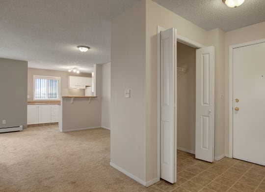 Empty apartment interior here at The Pointe at Applewood with beige carpet and tile flooring. Open closet and white front door on the right, leading to a bright kitchen with white cabinets.