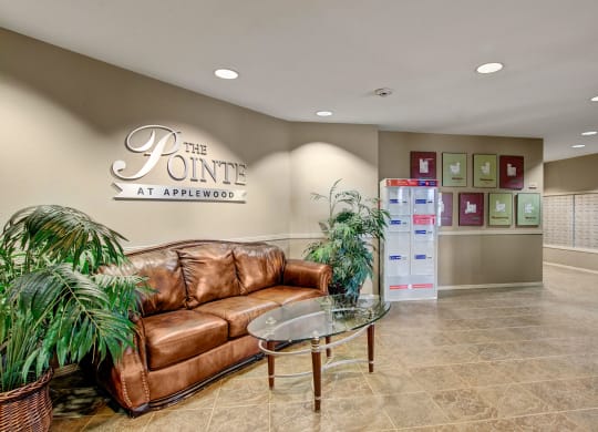 Cozy lobby with a brown leather couch, glass coffee table, and potted plants. Wall sign reads "The Pointe at Applewood." Mailboxes in background.