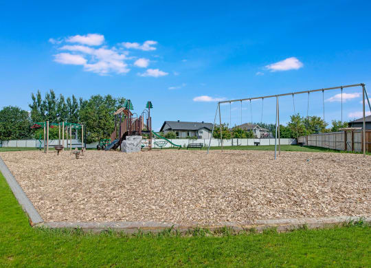 Playground here at Sol Terrace Apartment Homes with swings and climbing set, surrounded by wood chips and grass. Houses and trees in the background under a bright blue sky.