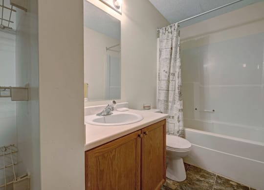 Small bathroom here at Sol Terrace Apartment Homes with wood cabinet, sink, and toilet. A white shower curtain with decorative patterns hangs over a bathtub. Light tones create a clean, simple look.
