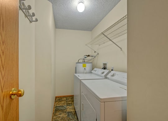 Laundry room here at Sol Terrace Apartment Homes with a washing machine, dryer, and water heater. An overhead shelf is mounted on the right wall. The room has beige walls and tiled floor.