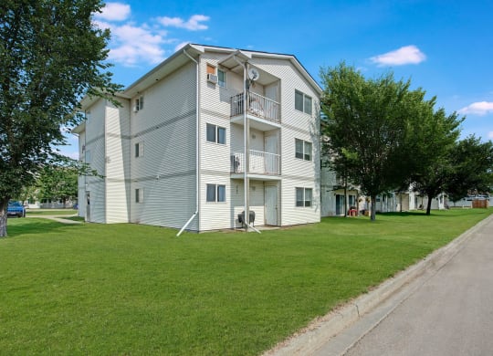 Three-story Sol Terrace apartment building with white siding, surrounded by green lawns and vibrant trees under a bright blue sky. A parked blue SUV is visible.