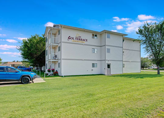 Four-story beige apartment building, labeled "Sol Terrace," with well-maintained lawn, trees, and a parked blue SUV under a bright blue sky.