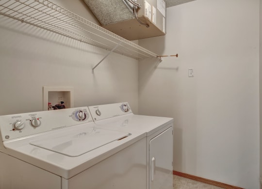 Small laundry room here at Villagio Apartment Homes with a white washer and dryer, white wire shelving above, and neutral walls. The space is clean and organized, conveying simplicity.