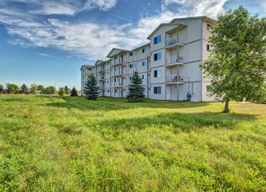 Four-story white Villagio Apartment Homes building with balconies on a sunny day, surrounded by a lush grassy lawn and trees. Blue sky with scattered clouds.