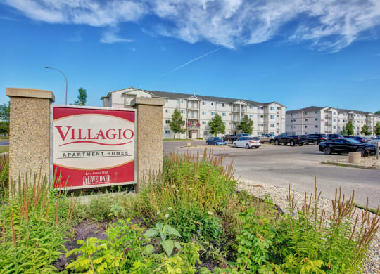 Sign reads "Villagio Apartment Homes" in front of a modern apartment complex with a parking lot and landscaped greenery under a clear blue sky.
