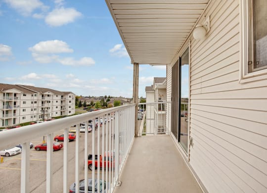 A beige apartment balcony here at Westhaven Estates Apartment Homes with white railings overlooks a parking lot and nearby buildings. The sky is blue with scattered clouds, creating a calm ambiance.