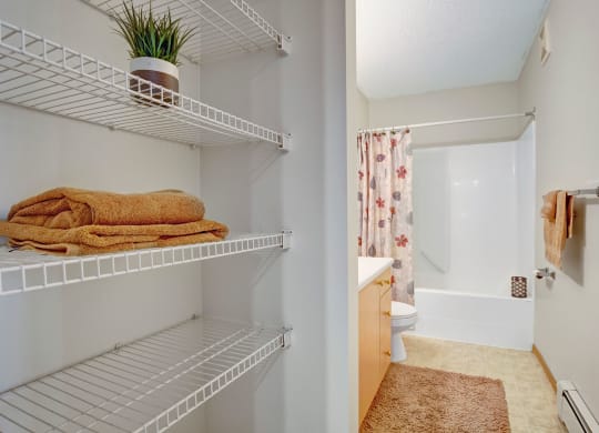 Neat bathroom here at Westhaven Estates Apartment Homes with floral curtain over tub. White shelves on the left hold folded towels and a plant. Soft, earth-toned decor creates a cozy vibe.