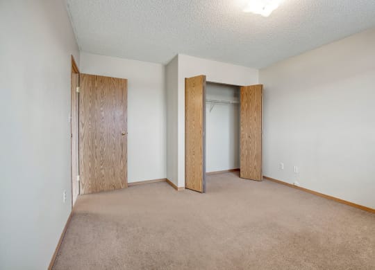 Empty room here at Westhaven Estates Apartment Homes with beige carpet, white walls, and a popcorn ceiling. Wooden door opens to the left, while a closet with bifold doors is on the right. Bright yet stark ambiance.