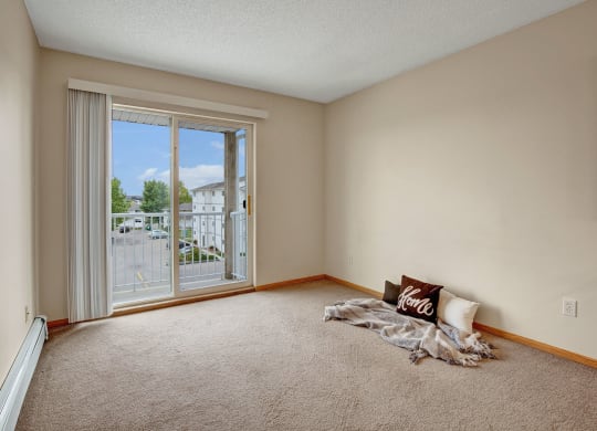 Empty room here at Westhaven Estates Apartment Homes with beige walls and carpet, featuring a sliding glass door leading to a balcony. A cozy corner has throw pillows and a blanket, conveying a welcoming feel.