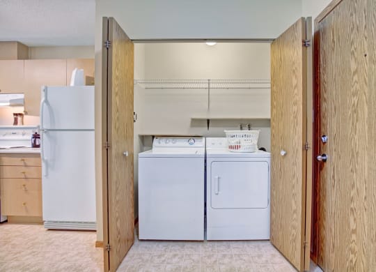 Laundry area here at Westhaven Estates Apartment Homes with wooden doors opened, revealing a white washer and dryer set. A wire shelf above holds a laundry basket. Adjacent kitchen features a fridge. Clean and organized space.