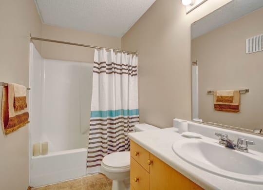 A clean, modern bathroom here at Westhaven Estates Apartment Homes with beige walls features a white bathtub and a striped shower curtain. A wooden vanity with a sink and a large mirror adds a tidy, fresh look.