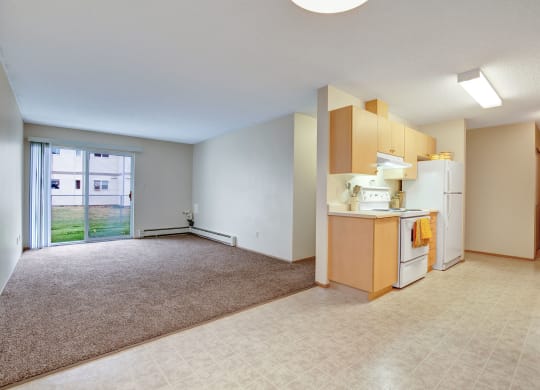 Open-plan apartment here at Westhaven Estates Apartment Homes with beige walls and mixed flooring. A sunny living area with large windows and a compact kitchen with wooden cabinets and white appliances.